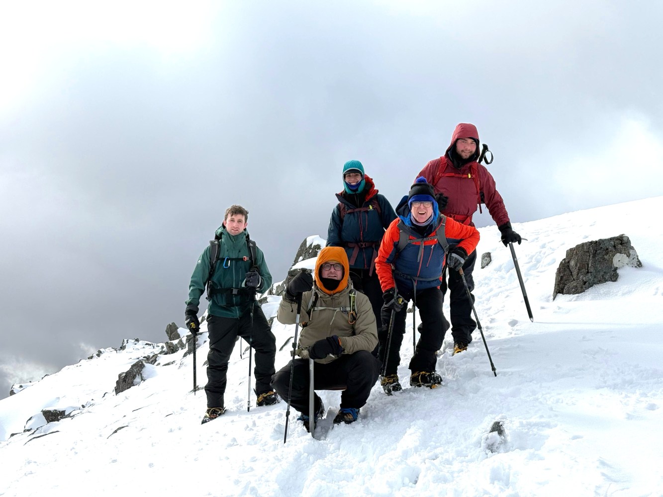 Five people in snow gear posing with hiking poles on a snowy mountain.
